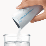 Hand die een zakje machine-ontkalker over een glas water giet, voor het reinigen van koffie- en theeapparatuur bij Koffie Thee Lokaal Delicious.