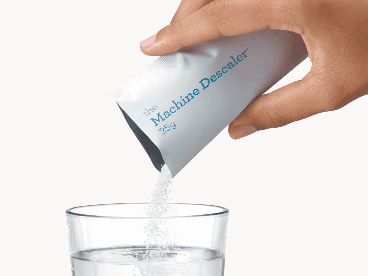 Hand die een zakje machine-ontkalker over een glas water giet, voor het reinigen van koffie- en theeapparatuur bij Koffie Thee Lokaal Delicious.