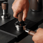 Zwarte espressomachine met hand die de koffiemolen bedient, in een koffie- en theelokaal, focus op versgemalen koffie, ambachtelijke koffiezetten, en koffie-ervaring.