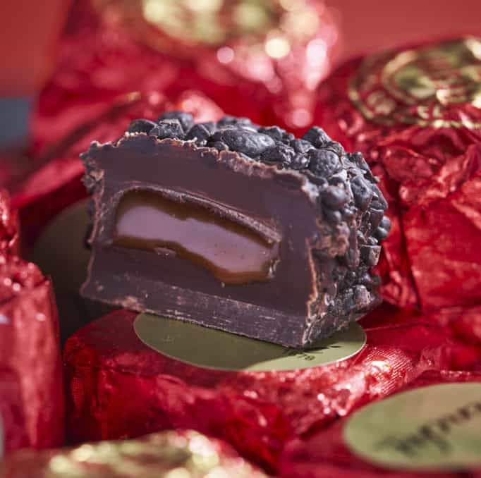 Luxe chocolade truffel met karamelvulling, omgeven door rood verpakkingspapier, perfect voor koffie en thee liefhebbers bij Koffie Thee Lokaal Delicious.