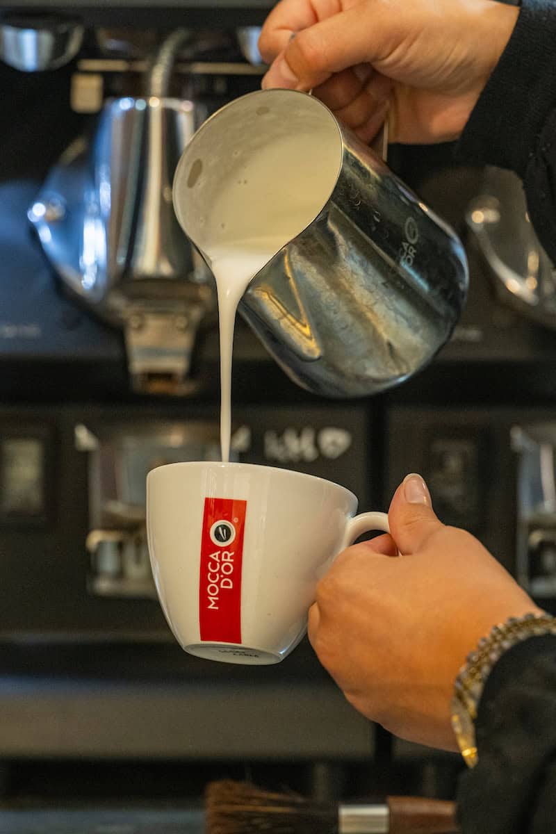 Koffie wordt ingeschonken in een wit kopje met rood logo, klaar voor een warme drank bij Koffie Thee Lokaal Delicious, de perfecte plek voor koffie- en theeliefhebbers in Nederland.